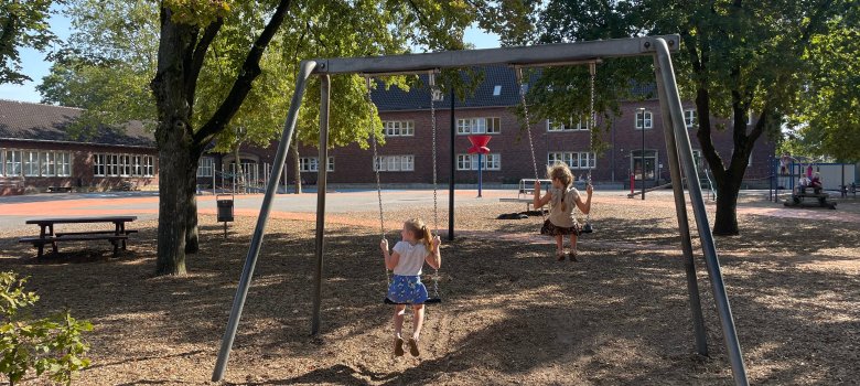 Het schoolplein met een schommel en zitje onder de bomen.